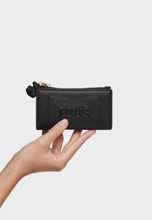 Mano sosteniendo una cartera de cuero negra con textura, con el logo "TOUS" en relieve y una cremallera dorada con un tirador en forma de oso negro.