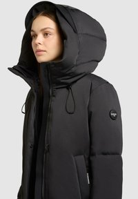 Zwarte gewatteerde jas met grote capuchon, ritssluiting en drukknoopsluiting; beschikt over elastische trekkoorden en een logopatch op de mouw.