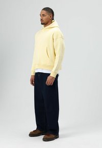 Gele capuchontrui met voorzak, gecombineerd met loszittende marineblauwe jeans en bruine veterlaarzen. Effen achtergrond voor betere zichtbaarheid.