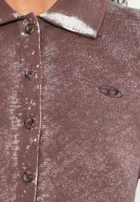 Detailný záber na hnedé pletené polo tričko so štyrmi gombíkmi, bielym škvrnným vzorom a malým čiernym vyšívaným logom na ľavej strane hrudníka.