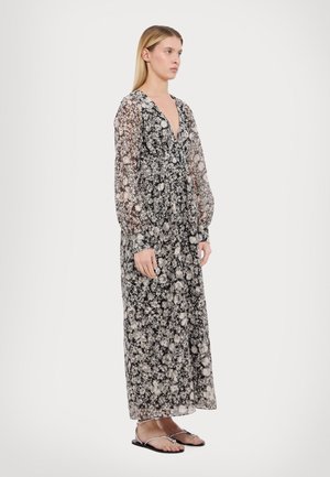 Maxi dress in sheer black fabric with a floral pattern. Features a deep V-neck and long sleeves, with a fitted waist. Worn with flat sandals.