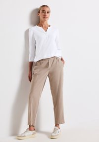 Frau, die an einer weißen Wand steht und ein weißes Langarm-Henley-Oberteil, beige Hose mit elastischem Bund und weiße Sneaker trägt.