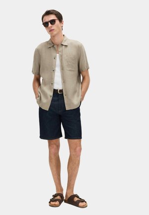 Muž v béžové krátkém košili na knoflíky přes bílý nátělník, tmavě modré džíny krátké kalhoty s černým pásem, hnědé sandály a černé sluneční brýle.