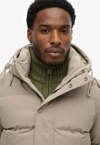 Beżowa puchowa kurtka z odpinanym kapturem, zapinana na zamek oraz ze sznureczkami. W okolicach kołnierza widoczny jest zielony wewnętrzny sweter z prążkowanego dzianego materiału.