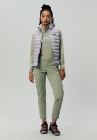 Gilet isolato grigio chiaro sopra una maglietta a maniche lunghe verde chiaro e pantaloni aderenti. Scarpe da escursionismo nere e rosa con calzini rosa. Texture lisce ovunque.
