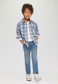 Camicia a quadri blu e verde con bottoni, t-shirt bianca sotto, jeans azzurri chiari e sneakers bianche con dettagli grigi. Capelli texturizzati.