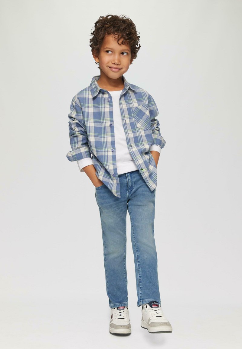 Camicia a quadri blu e verde con bottoni, t-shirt bianca sotto, jeans azzurri chiari e sneakers bianche con dettagli grigi. Capelli texturizzati.