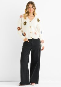 Knit cardigan with a deep V-neck, cream color, featuring colorful floral appliqués, and wooden buttons. Paired with wide-legged black jeans.