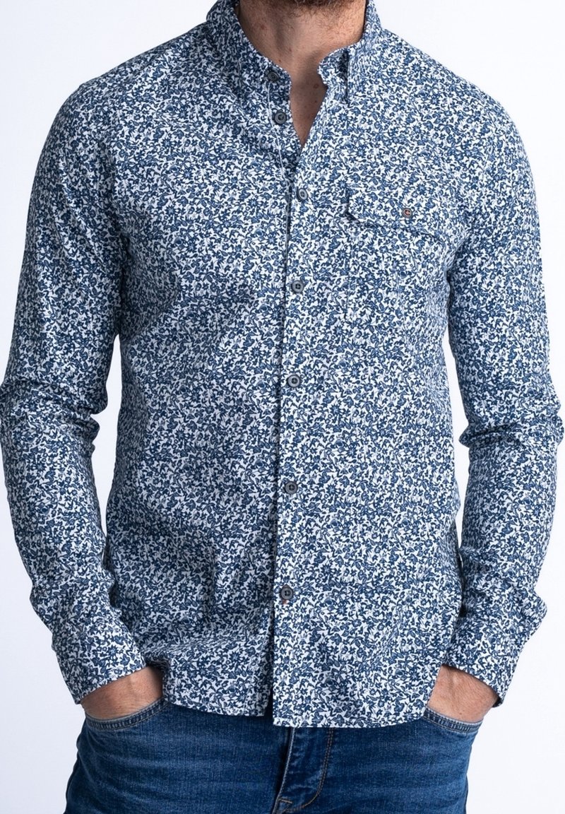 Homme portant une chemise blanche à manches longues avec un motif floral bleu, boutonnée sur le devant, poche poitrine, mains dans les poches d'un jean bleu.