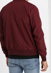 Chaqueta bomber burdeos con cinturilla acanalada. Tela suave y brillante con puños elásticos, combinada con jeans de color azul claro. Sin logotipos visibles.