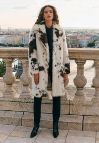 Umělá kožešinová bunda s hnědo-krémovým skvrnitým vzorem, s širokým límcem a zapínáním na knoflíky, v kombinaci s černými upnutými kalhotami a špičatými botami.