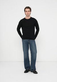 Schwarzes Langarmshirt kombiniert mit locker sitzenden blauen Jeans. Das Modell steht mit den Händen in den Taschen, trägt schwarze Schuhe vor einem weißen Hintergrund.