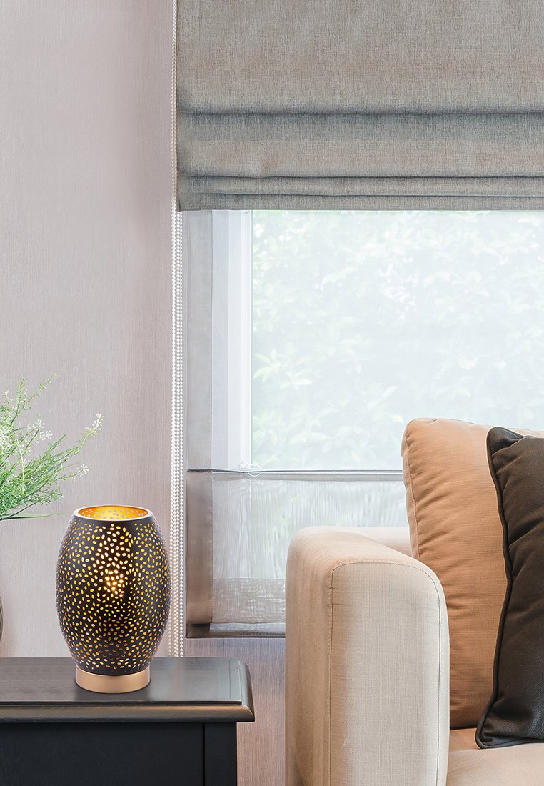 Lampe en céramique noire et or avec un design perforé, reposant sur une table en bois foncé à côté d'un canapé beige et d'un rideau léger.