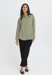 Giovane donna che sorride, indossando una camicia a righe verde e bianca con bottoni, pantaloni neri e scarpe slingback nere a punta, in piedi con le gambe incrociate.