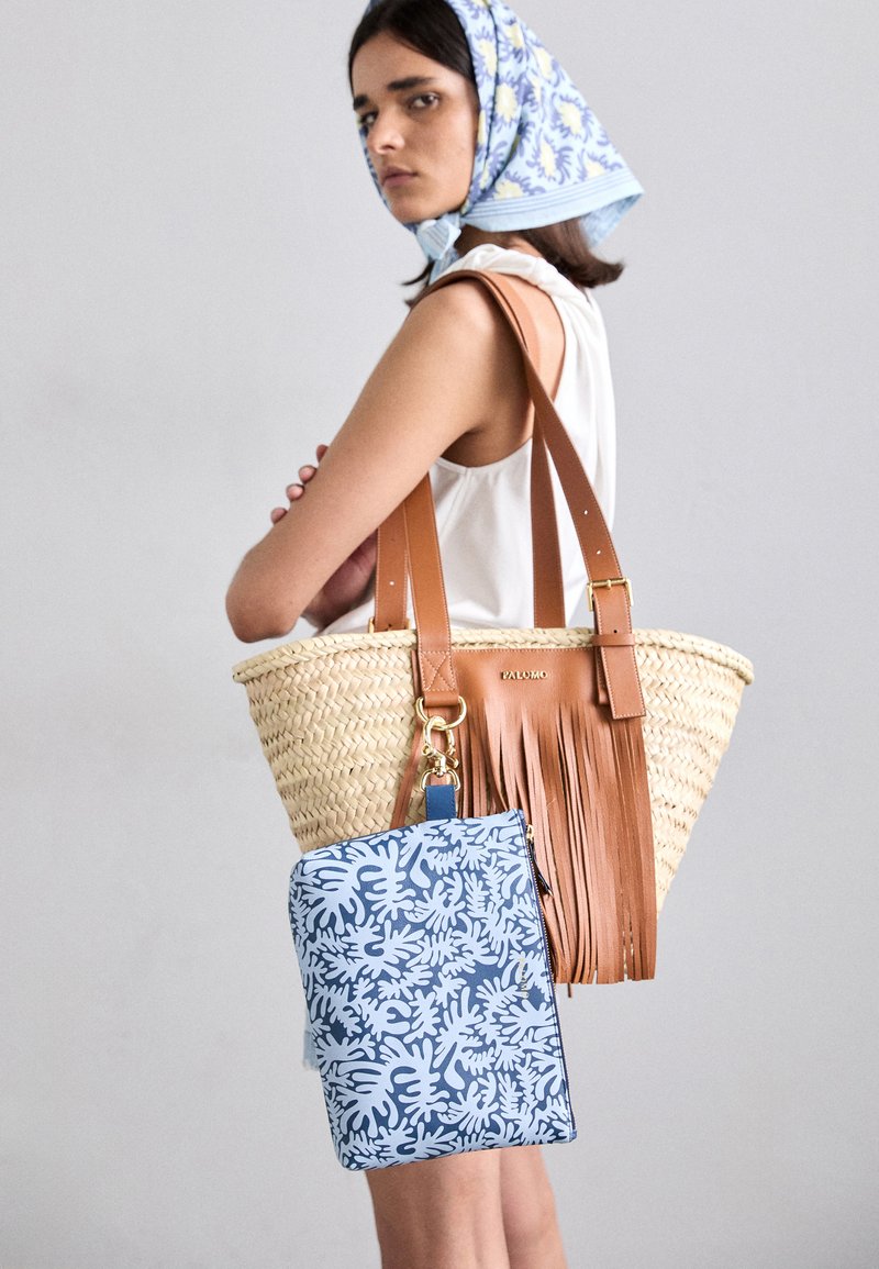 Woven straw tote bag with brown leather straps and fringe detail, paired with a blue floral-patterned zippered pouch.