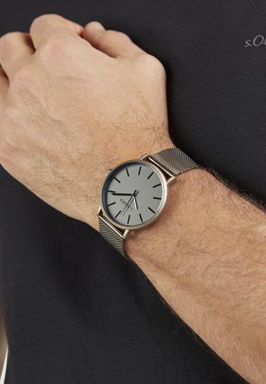 Zilverkleurige horlogeband van mesh met een ronde grijze wijzerplaat met dunne uurmarkeringen en een minimalistisch ontwerp, gedragen om een pols tegen een zwarte stoffen achtergrond.