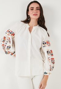 Witte katoenen blouse met pofmouwen, voorzien van kleurrijke bloemenborduursels op de manchetten en schouders, en een knoopsluiting aan de voorkant.
