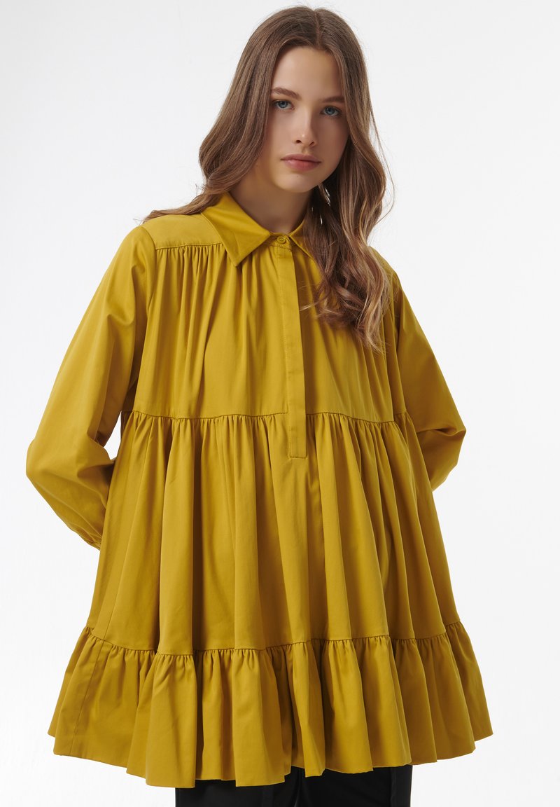 Mosterdgele katoenen blouse met een gerimpelde zoom, lange mouwen en een kraaghals. Heeft geplooide details onder de borst.