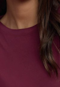 Chemise bordeaux avec une texture lisse, un col rond, et des détails de couture subtils, complétant des cheveux lâches et ondulés au premier plan.