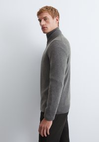 Gray, high-collar sweater in a textured knit, with long sleeves and a slim fit, paired with dark pants, viewed from the side.