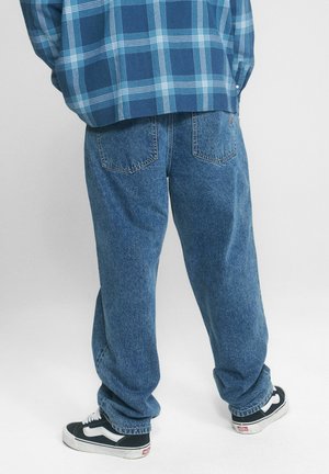 Osoba nosząca luźne niebieskie dżinsy, niebieską koszulę w kratę oraz czarno-białe tenisówki stojąca na jednolitym jasnym tle.