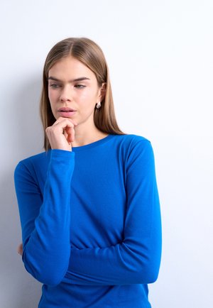 Jeune femme en chemise bleue à manches longues, aux longs cheveux, appuyant le menton sur la main, regardant pensivement sur le côté sur fond blanc.