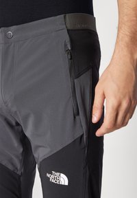 Muška ruka blizu zatvorenog bočnog džepa crno-sivih hlača North Face s logom na butini.