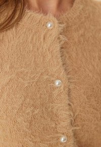 Detailní záběr na huňatý béžový pletený kardigan se třemi perlovými knoflíky zapnutými na přední straně, umístěný na horní části hrudníku osoby.