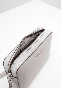 Šedá syntetická crossbody taška se zipovým uzávěrem, hladkým povrchem a řetízkovým popruhem. Obsahuje logo detail na přední straně.