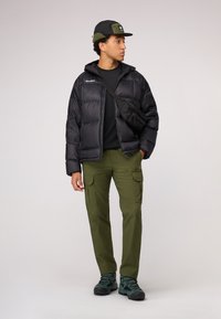 Une doudoune noire, un pantalon cargo vert olive, un sac noir, une chemise sombre, une casquette noire avec des accents verts et des bottes de randonnée sombres, se tenant sur un fond clair.