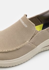 Beige liukastuvasta kenkä, jossa on teksturoitu verkkopinta, elastiset sivupaneelit ja kumipohja. Sivussa on valkoinen logo.