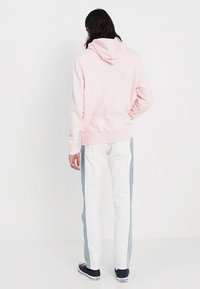 Sudadera rosa claro hecha de tela suave con capucha, combinada con jeans blancos que tienen paneles laterales azul claro. Zapatillas completan el conjunto.