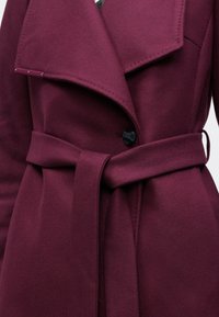 Bourgogne ullrock med stor krage, bältad midja och en enda svart knappstängning. Har en mjuk textur och snygga sömdetaljer.