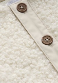 Close-up van crèmekleurige boucle stof met een beige stofstrip en twee bruine viergaatjesknopen genaaid.