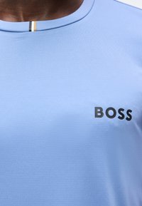 Κοντινή λήψη ενός ανοιχτό μπλε μπλουζάκι με στρογγυλή λαιμόκοψη και το λογότυπο "BOSS" τυπωμένο με μαύρο χρώμα στο αριστερό μέρος του στήθους, καθώς και μια μικρή λευκή λεπτομέρεια με ρίγες στο εσωτερικό του γιακά.