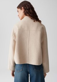 Donna con capelli castani ondulati, che indossa una giacca di lana beige e jeans blu, rivolta di spalle su uno sfondo grigio chiaro uniforme.