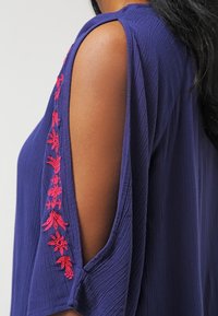 Mouwdetail van een paarse blouse met open schouder en verticale rode bloemenborduursels langs de uitsnijding.