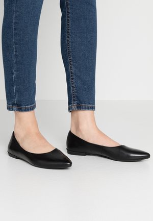 Person trägt dunkelblaue Skinny-Jeans und schwarze flache Schuhe mit spitz zulaufender Vorderseite, steht auf einem hellgrauen Boden.