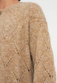 Κοντινό πλάνο ενός ατόμου που φοράει ένα απαλό πλεκτό πουλόβερ σε καμηλό χρώμα, με διαμάντι σχέδιο και λεπτές διακοσμητικές διατρήσεις.