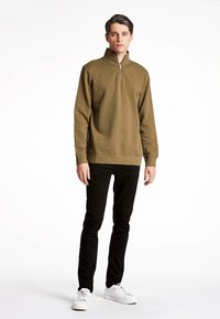 Maglione verde oliva con colletto a mezza zip, texture morbida e dettaglio circolare ricamato sul petto, abbinato a pantaloni neri e sneakers bianche.