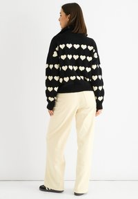 Zwarte gebreide trui met een oversized pasvorm, voorzien van crème hartpatronen. Gecombineerd met losse beige broek en zwarte sneakers.