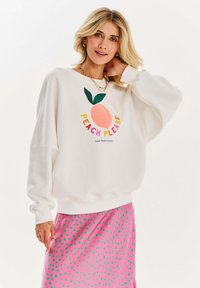 Bílá mikina s grafikou "Peach Please", která zobrazuje ilustraci broskve s zeleným listem. Spárováno s růžovou sukní s zelenými puntíky.