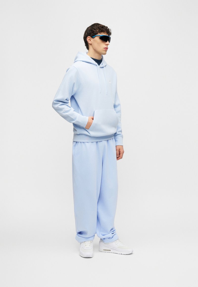 Joven con sudadera con capucha azul claro y pantalones deportivos a juego, zapatillas blancas y gafas de sol oscuras envolventes, de pie con una mano en el bolsillo.