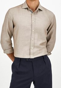 Camicia in lino marrone chiaro con maniche arrotolate, design con bottoni e tessuto sottile. Abbinata a pantaloni sartoriali blu scuro.