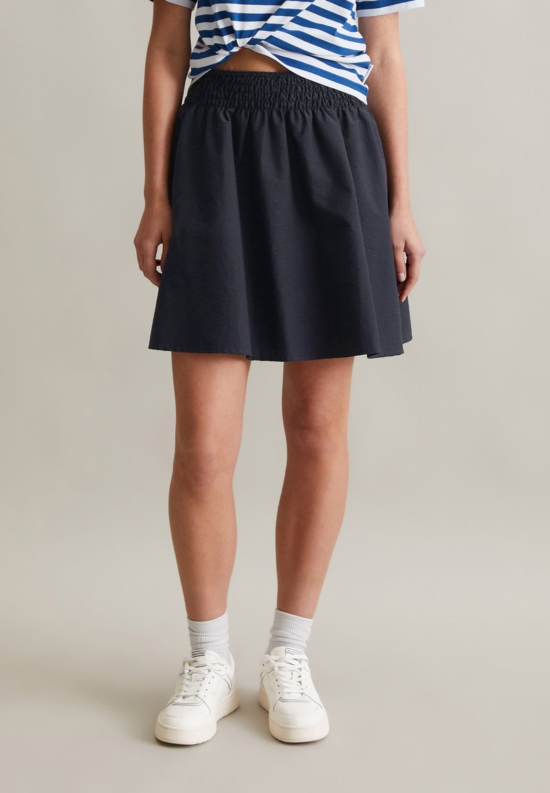 Zwarte rok met een elastische tailleband, A-lijn silhouet, gladde textuur en knielange afwerking, gedragen met witte sneakers en sokken.