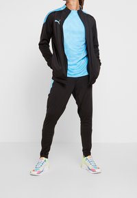 Schwarzer sportlicher Trainingsanzug mit blauen Akzenten, bestehend aus einer Jacke mit Reißverschluss und schmal zulaufenden Hosen. Leichtes, atmungsaktives Material. Bunte Sneakers.