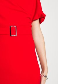 Robe rouge à manches courtes, dotée d'une boucle rectangulaire argentée à la taille. Texture lisse et ajustée avec des lignes épurées.