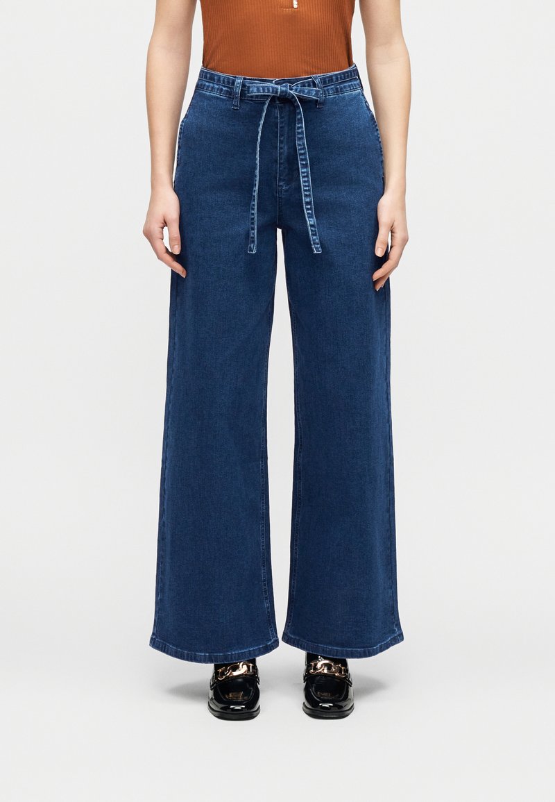 Γυναίκα που φοράει μπλε τζιν παντελόνι με φαρδιά μπατζάκια και ψηλή μέση, με υφασμάτινη ζώνη, σε συνδυασμό με αμάνικο τοπ σε σκουριά απόχρωση και μαύρα loafers με χρυσή αλυσίδα.