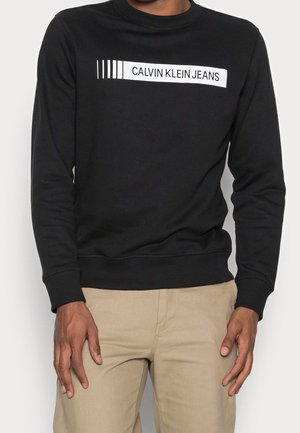 Mężczyzna w czarnej bluzie z napisem "CALVIN KLEIN JEANS" na piersi oraz beżowych spodniach, stojący na jednolitym tle.