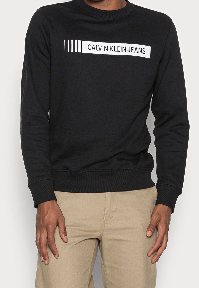 Homme portant un sweat-shirt noir avec le texte « CALVIN KLEIN JEANS » sur la poitrine et un pantalon beige, debout devant un fond uni.